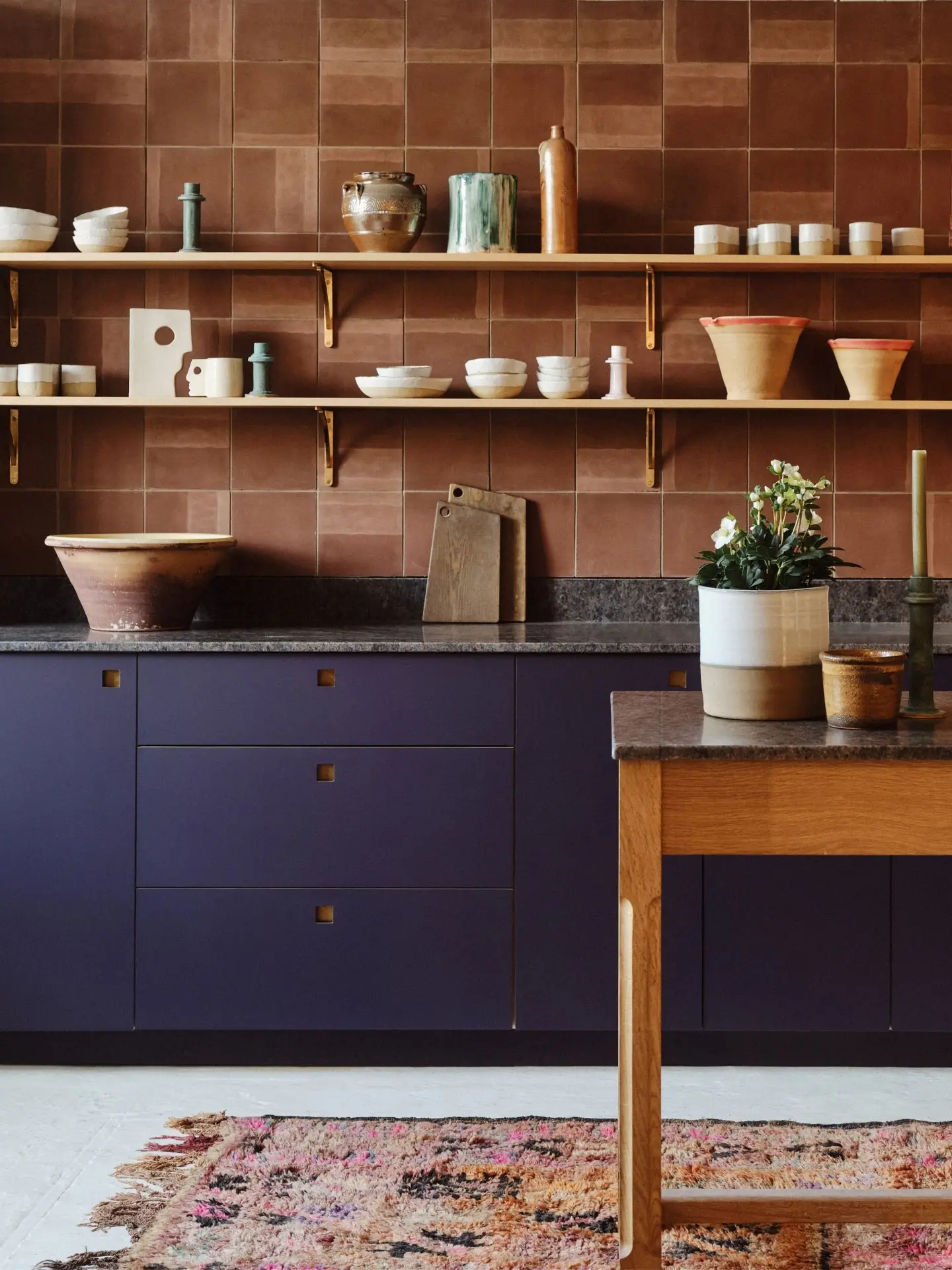 Stylish kitchen with aubergine colour cabinets, brown tiled walls, wooden shelves holding ceramics, and a cosy vintage rug.