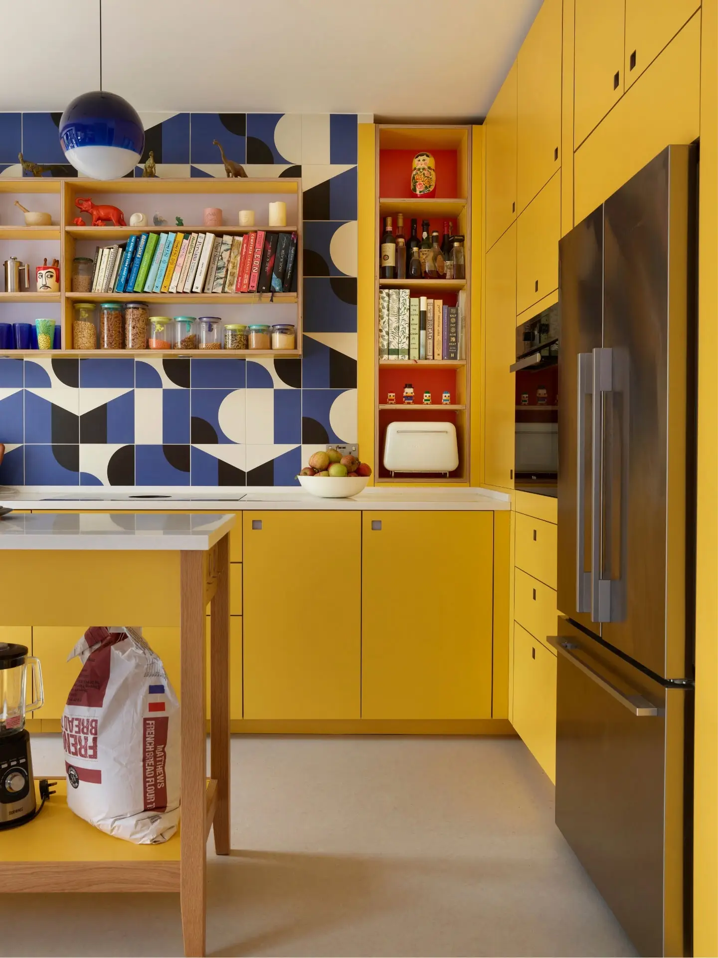 Bright kitchen with bold yellow cabinets, blue geometric tiled backsplash, and open shelves with books and jars.
