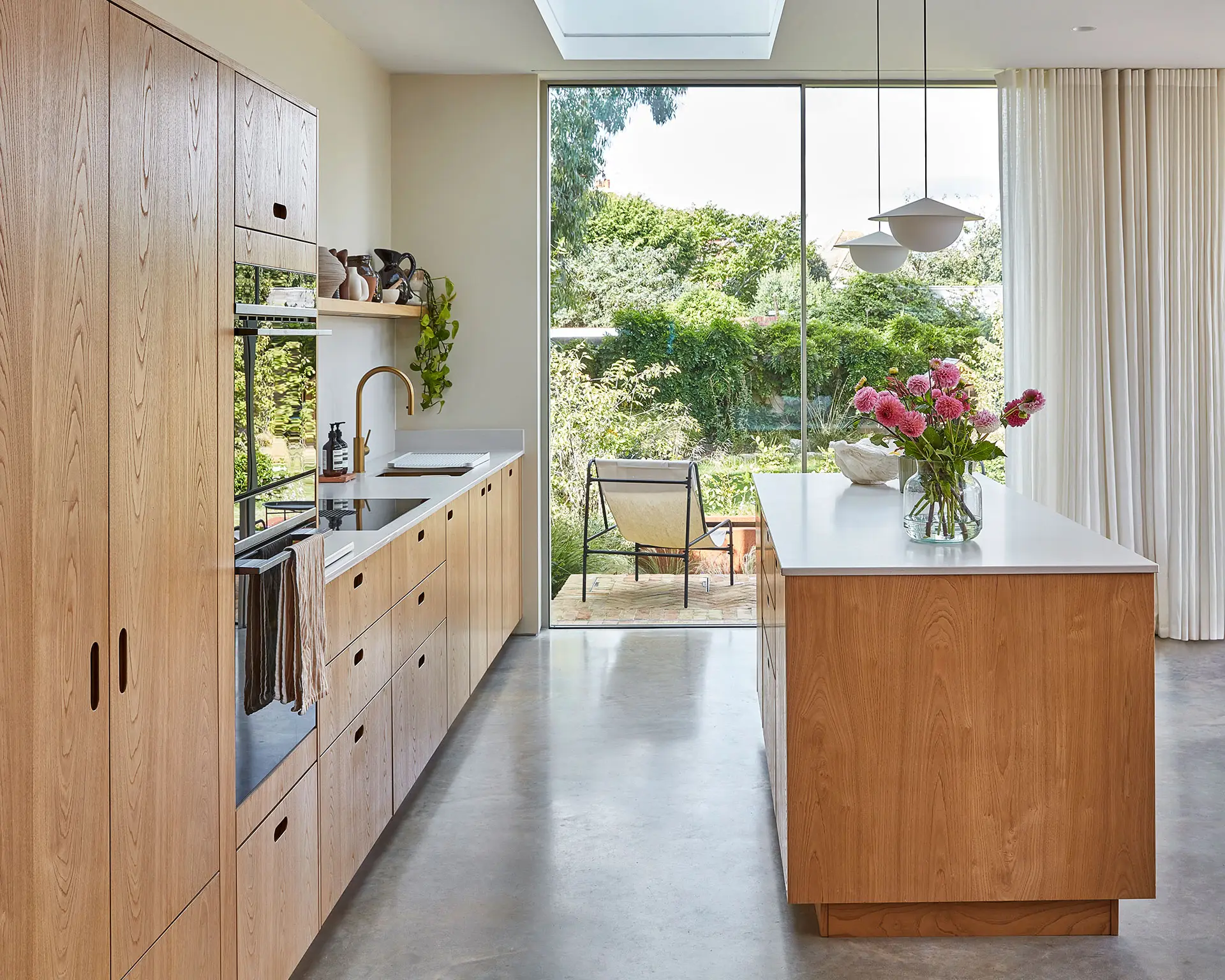 Pluck-Journal-The-Scandi-Chic-Kitchen-3 A kitchen in a family home in Brighton with wood cabinetry and brass handles, including an island, larder and shelf and grey concrete floor.