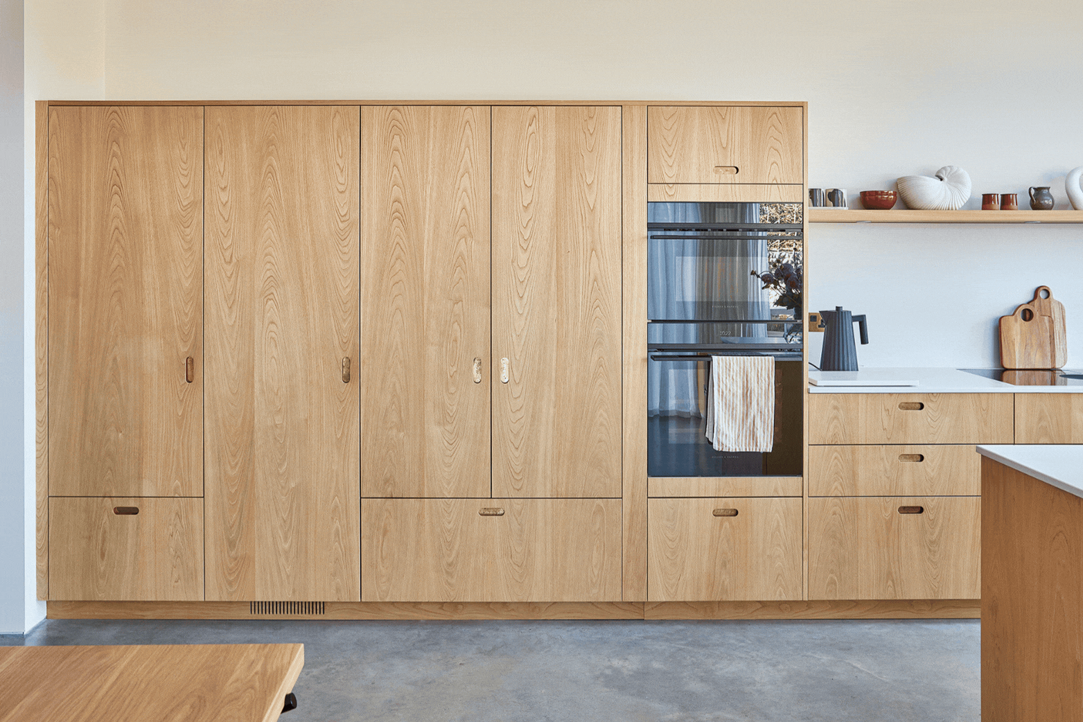 Pluck The Scandi Chic Kitchen GIF A kitchen in a family home in Brighton with wood cabinetry and brass handles, including an island, larder and shelf and grey concrete floor.