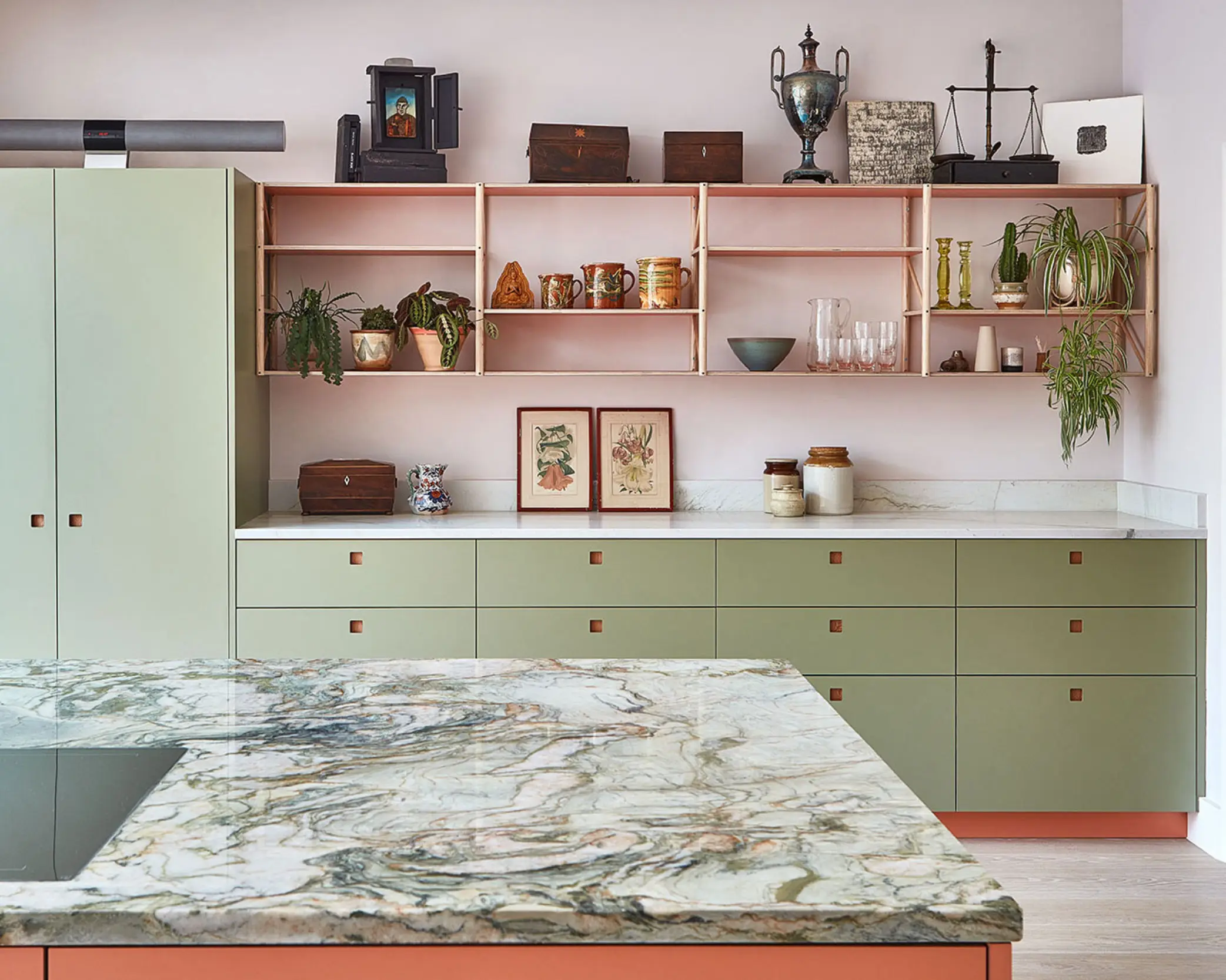 Green kitchen cabinetry with a coral pink island topped with swirling natural stone quartzite and a dining table.